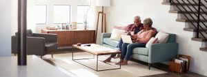 A retired couple sitting on a green sofa in a modern living room looking at a laptop and drinking tea