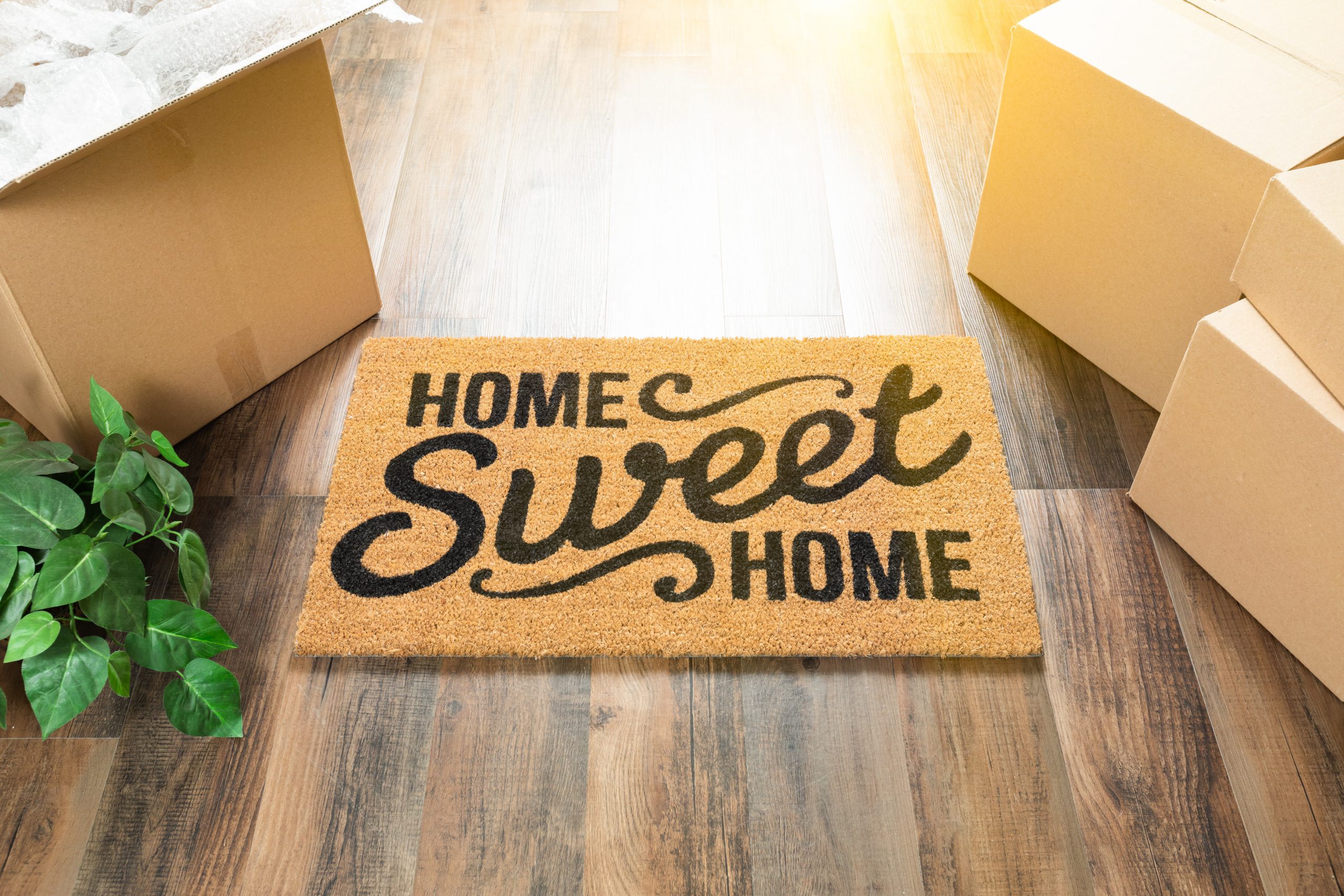 Front door mat with ‘home sweet home’ written on it, on a wooden floor and boxes around it