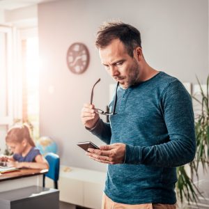 First time buyer father looking at smart phone with eyeglasses in his hand, in background his daughter doing homework, looking at houses for sale