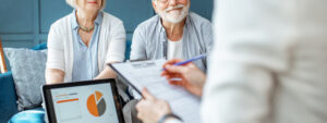 Senior couple on sofa at home facing a financial adviser with laptop and clipboard, talking about financial worries