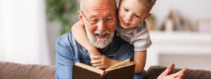 A grandfather reading a story from a book to his grandson on the sofa, retirement today
