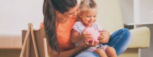 Mother sitting on floor with young daughter, hands around a piggy bank, clear goals for investment