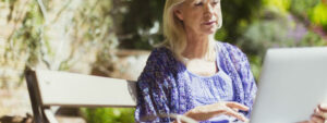 Senior woman using laptop on sunny garden bench, checking how age affects life insurance