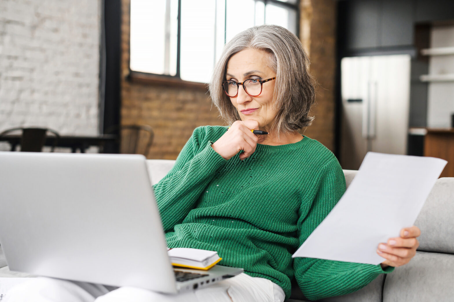 A mature, senior lady looking at laptop on sofa at home, with document in hand, working out her equity release cash