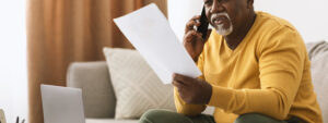 Senior Black Businessman Talking On Mobile Phone Holding Papers Working Sitting At Laptop Indoors. Mature African American Man Speaking By Cellphone In Living Room At Home. Communication Concept