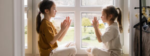 Two young girls sit on a windowsill, engaged in a playful game of clapping hands. The image captures a moment of innocent fun and joyful interaction between sisters or friends. The natural light streaming in adds warmth and creates a cheerful atmosphere.